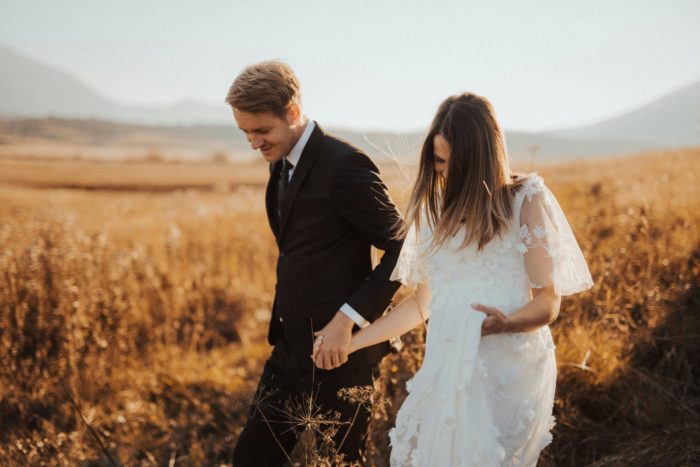 A bride in a delicate lace wedding dress and groom in a classic black suit walking together through a golden field at sunset, symbolizing the journey of marriage and the importance of discussing prenuptial agreements before taking this significant step together.