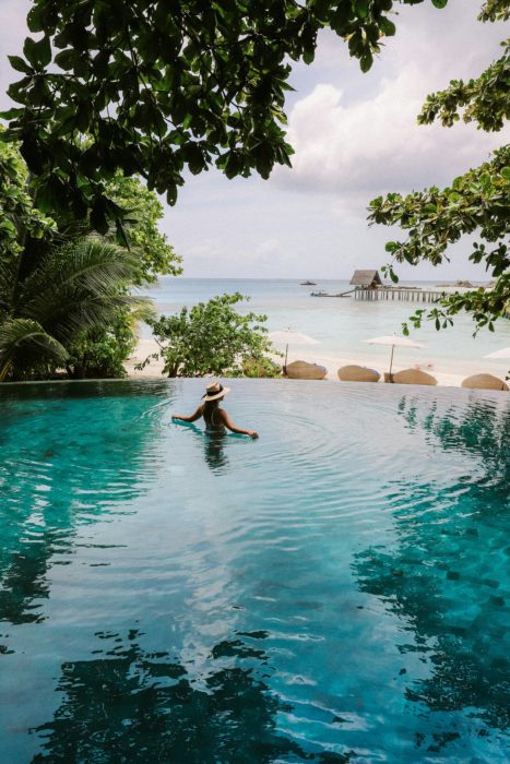 The image shows a person wearing a straw hat relaxing in an infinity pool with turquoise water that appears to blend seamlessly with the ocean beyond. The swimming pool is surrounded by lush tropical foliage, with palm trees framing the view. In the distance, there's a beach with lounge chairs and umbrellas, and a traditional over-water bungalow structure built on stilts extending into the clear blue sea. This serene tropical resort setting illustrates the concept of "habitual residence" mentioned in the blog post about international divorces, where couples may have connections to multiple countries through residences in exotic locations. Such luxury international properties could be among the assets that might be subject to different rules of division depending on which country's jurisdiction applies to divorce proceedings.