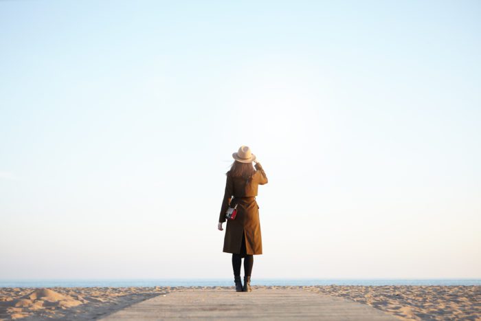 A woman in a coat and hat stands on a boardwalk looking out at a calm sea and a vast, open sky, symbolising a hopeful and peaceful future.