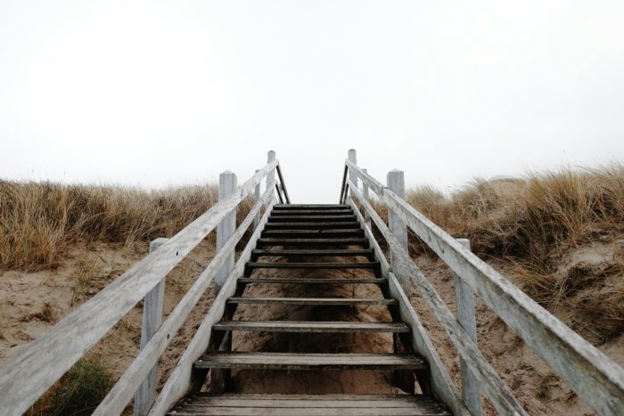 A set of weathered wooden stairs leading upwards through marram grass sand dunes towards a bright, misty horizon, symbolising a new path or chapter.